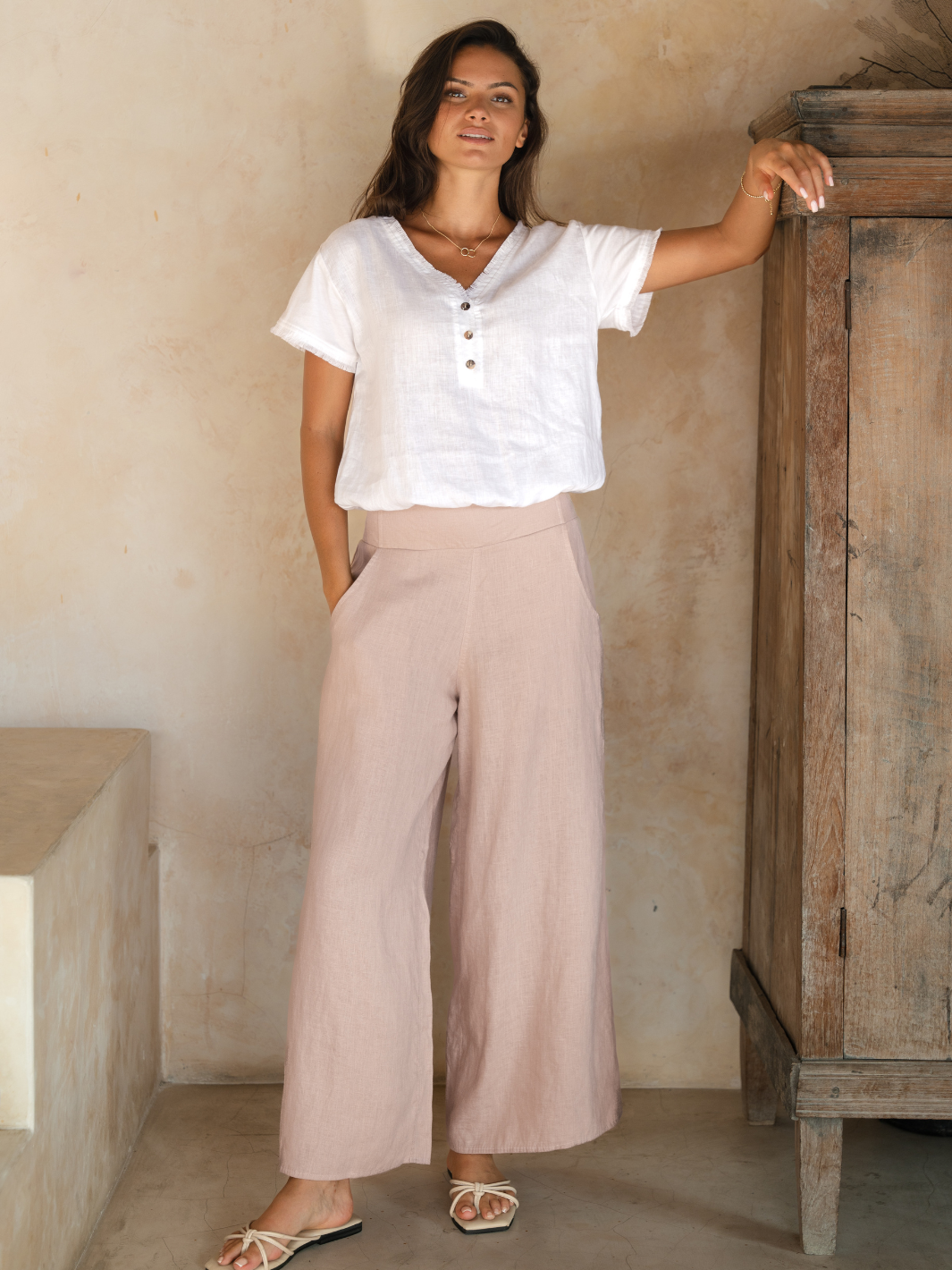 A woman standing in front of a rustic door, wearing a white top and pink wide-leg linen pants with a flared design.