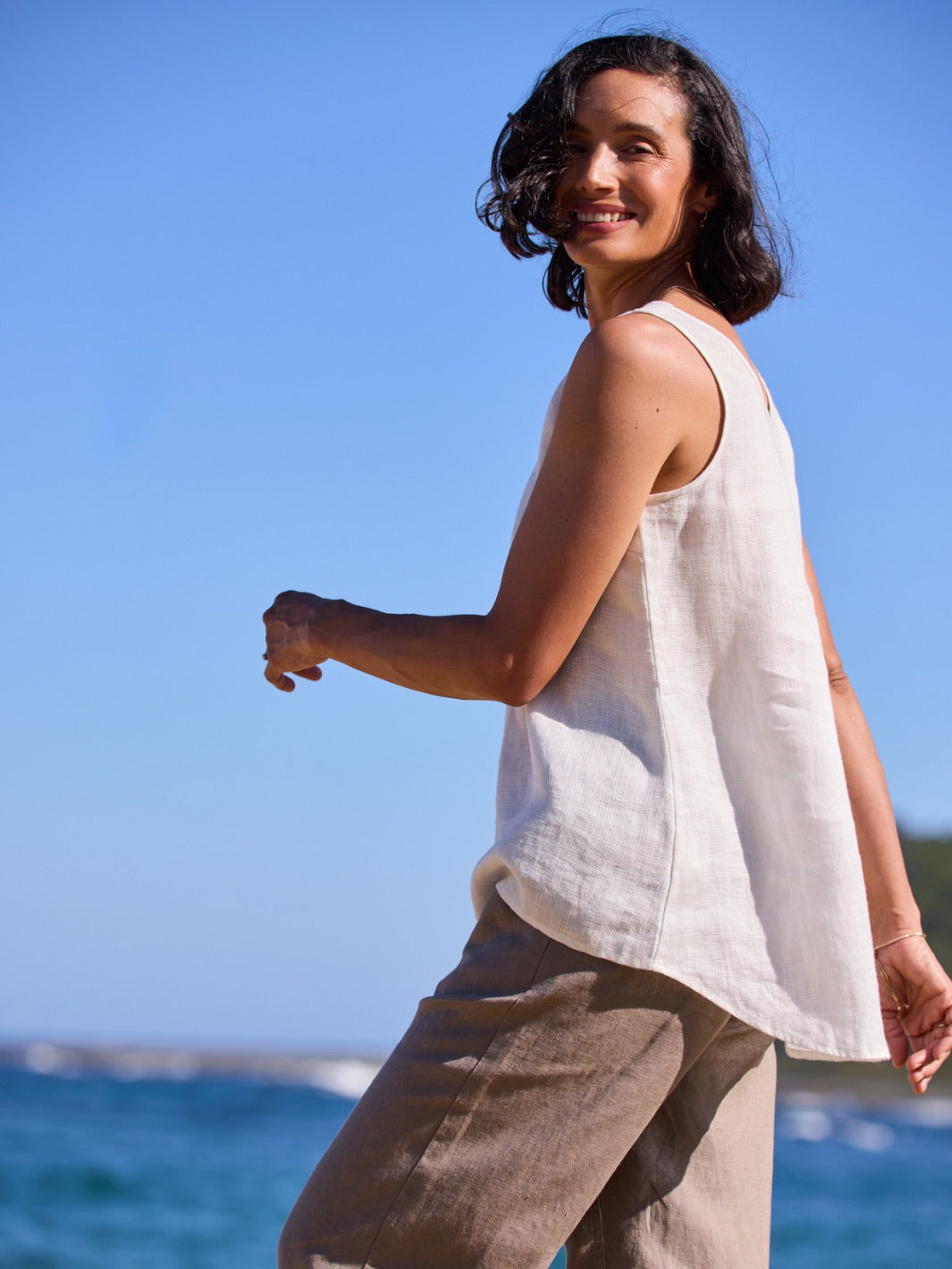 Woman standing on a beach wearing a sleeveless top and pants with a clear blue sky in the background.