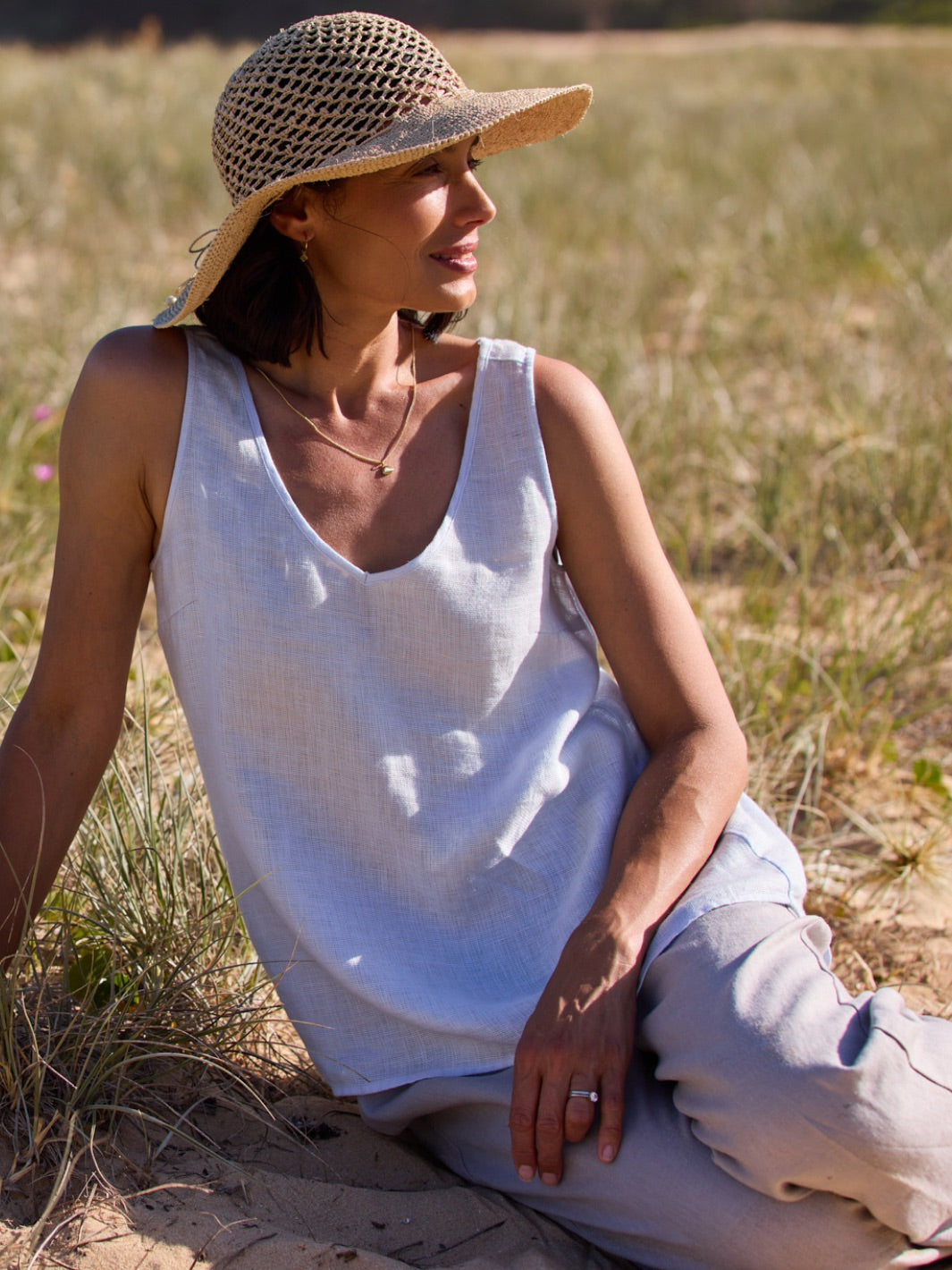 Woman sitting in a field wearing a white tank top and straw hat.