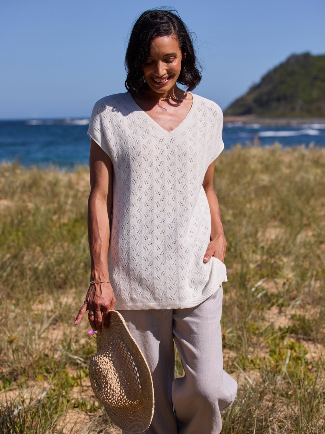 Woman standing on a grassy beach holding a hat, with ocean and sky in the background