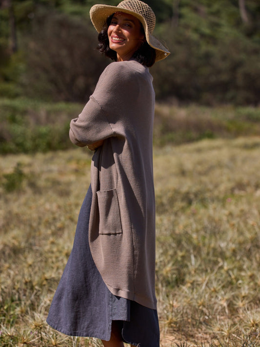 Woman wearing a long brown cardigan over a blue dress, standing in a natural setting with greenery.