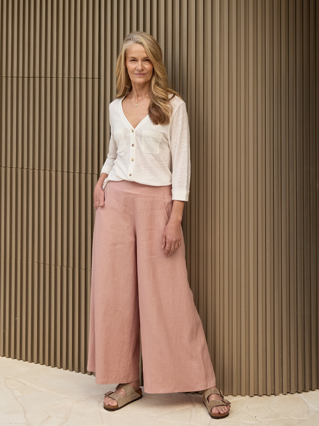 Woman wearing a white blouse and pink pants standing against a brown wall.
