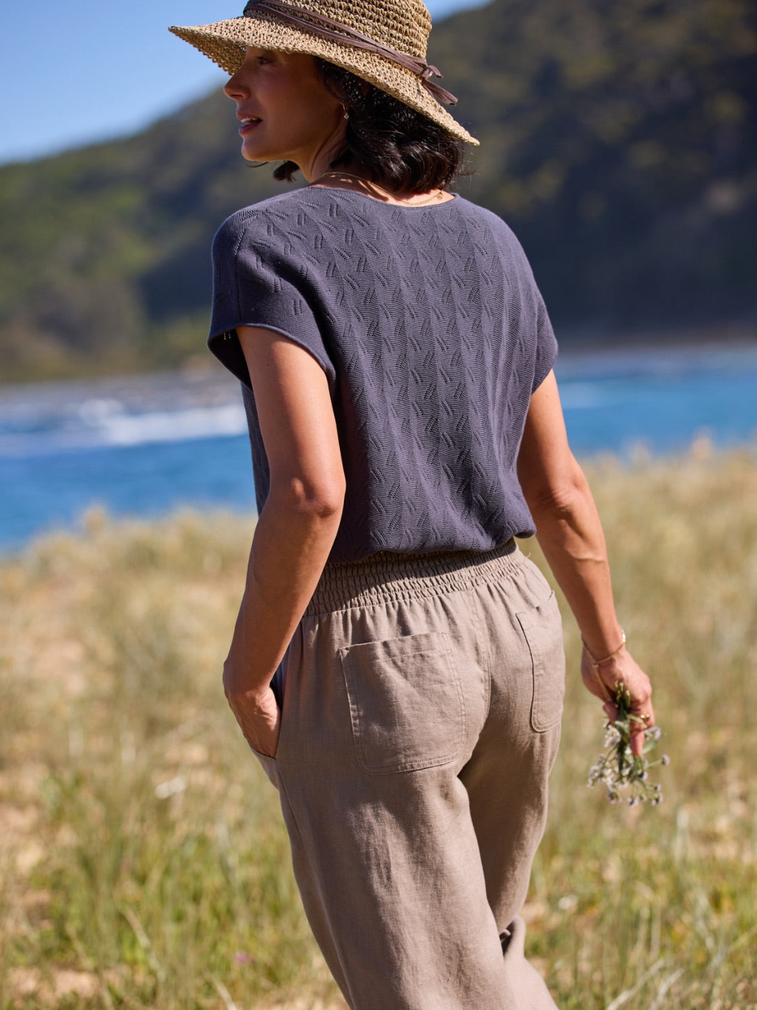Person wearing a dark knitted top and beige pants, standing in a natural setting with mountains and water in the background.