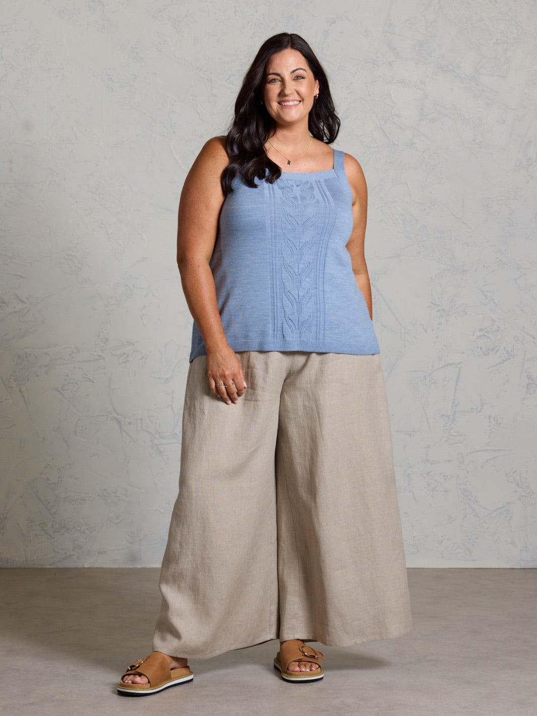 Woman wearing a blue sleeveless top and beige wide-leg pants against a gray background