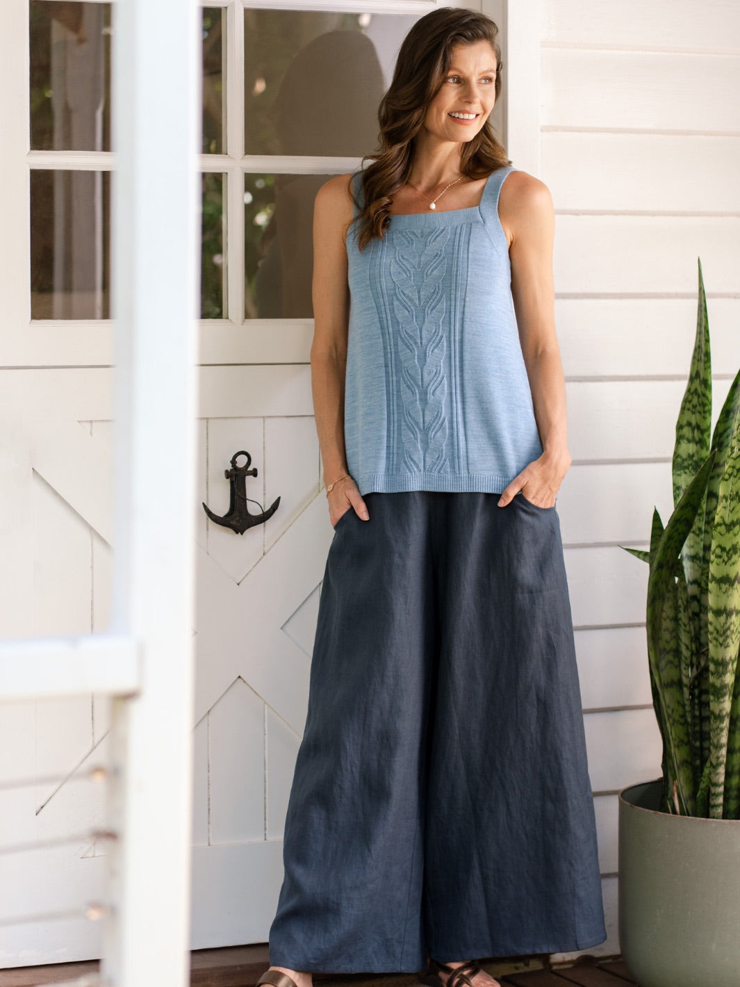 Woman wearing a blue sleeveless top and dark wide-leg pants standing on a porch.