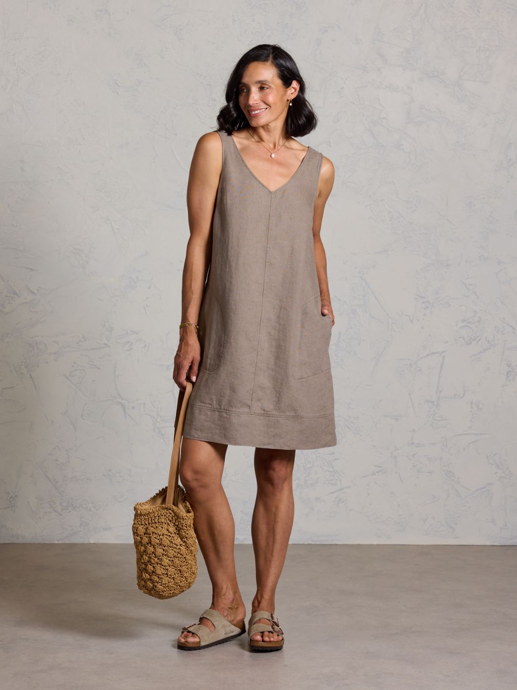 Woman wearing a brown sleeveless dress holding a woven bag against a plain background