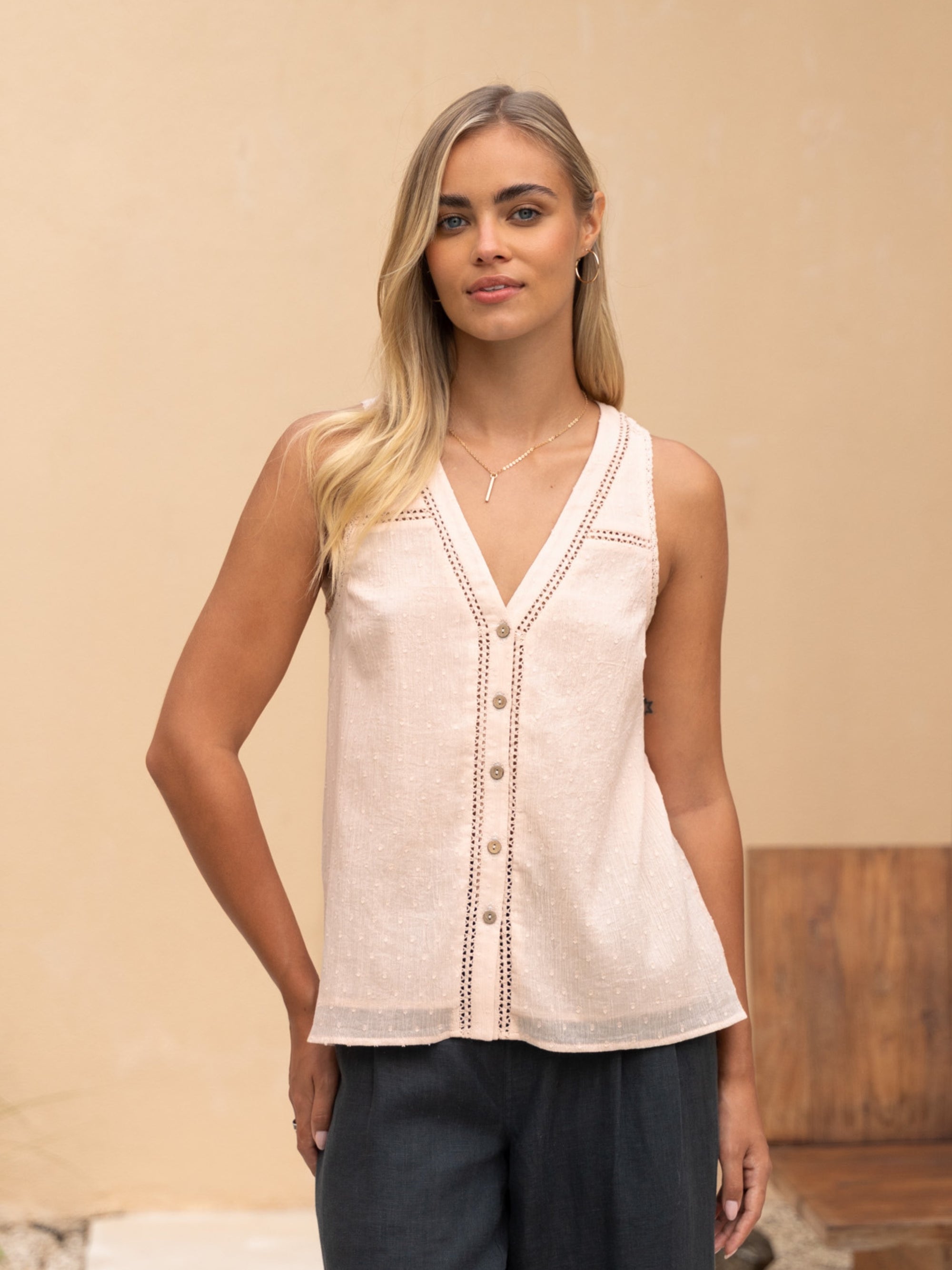 A woman wearing a beige cotton top with button details and lace trim, paired with dark pants.