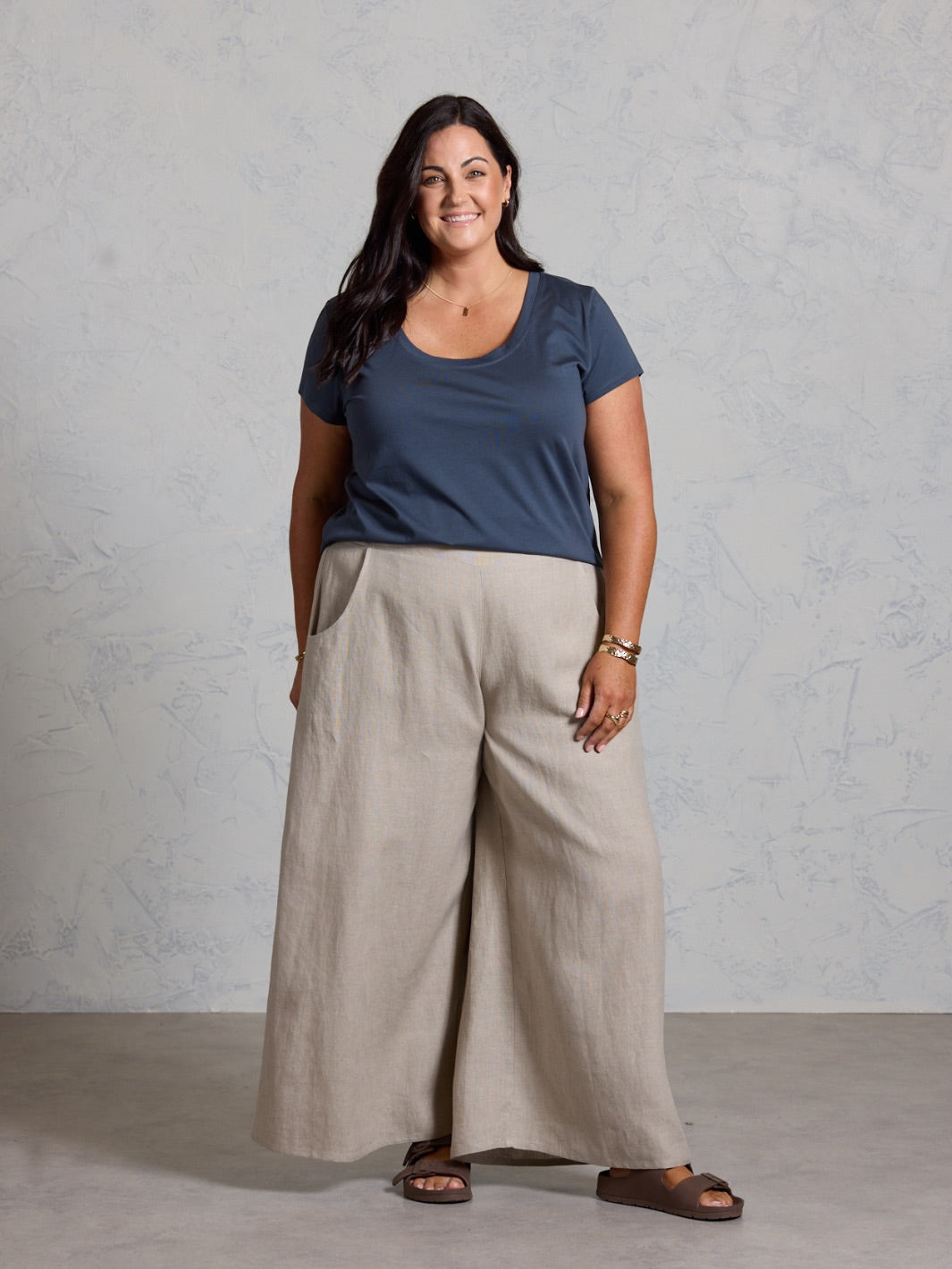 Woman wearing a black top and beige pants against a gray background