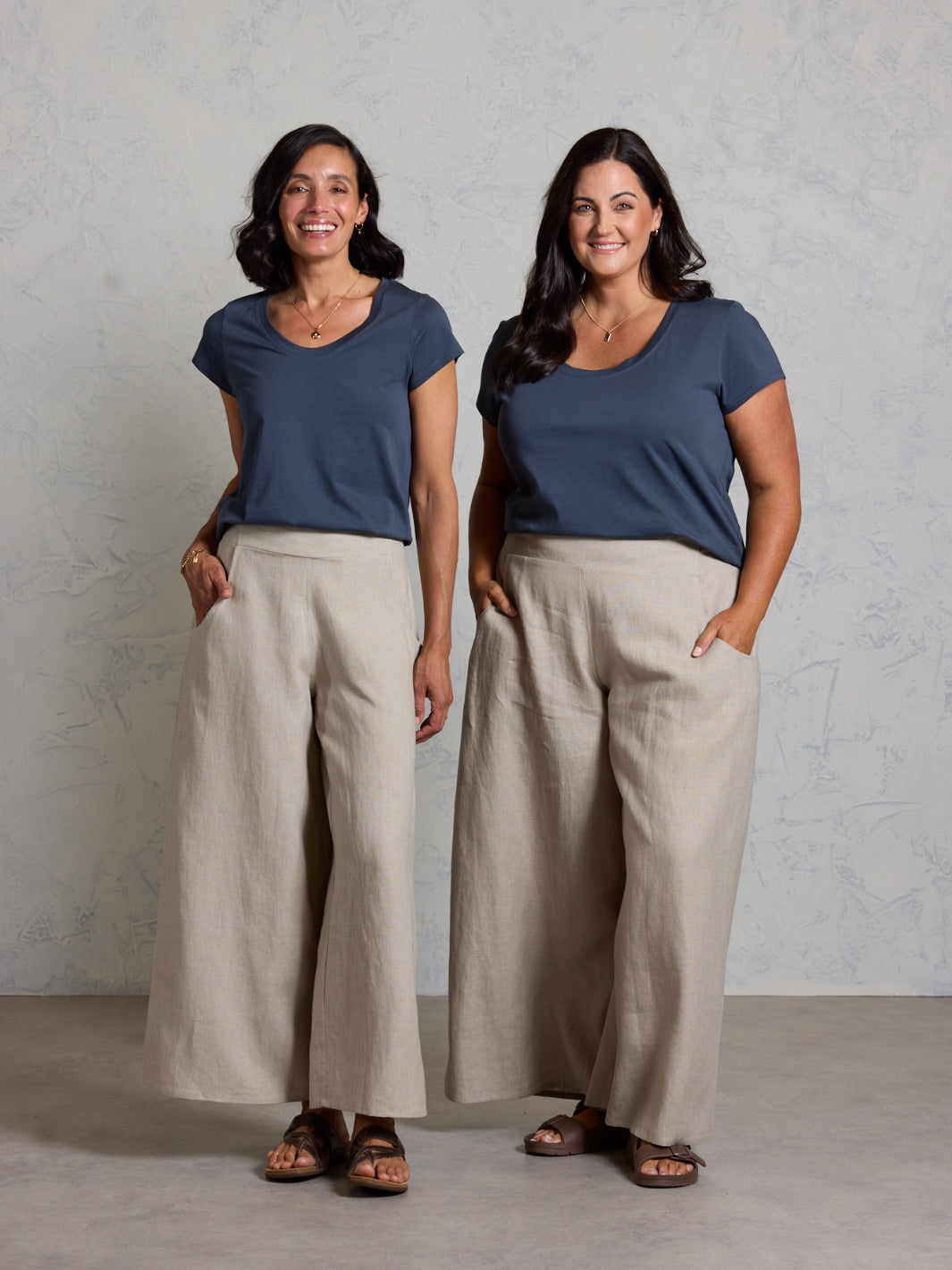 Two women wearing matching outfits of black tops and beige pants against a plain background.