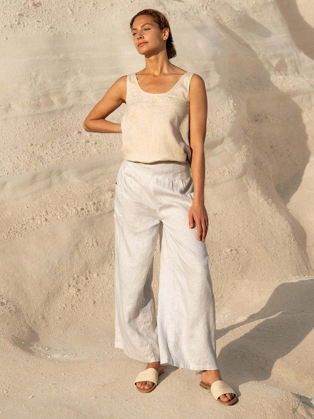 Woman in a beige tank top and light-colored pants standing on sandy ground.