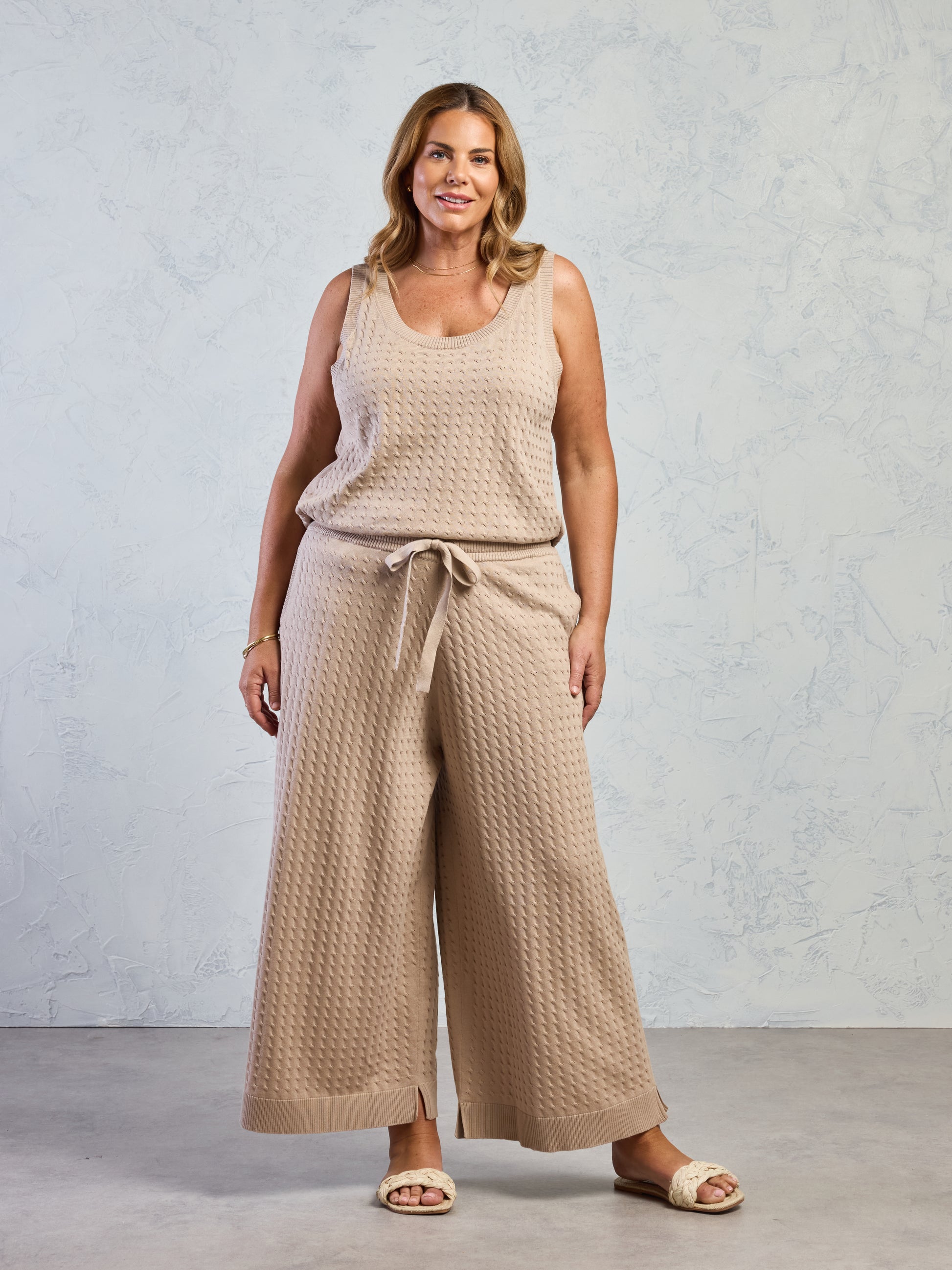 Woman wearing a beige sleeveless top and wide-leg pants against a light gray background