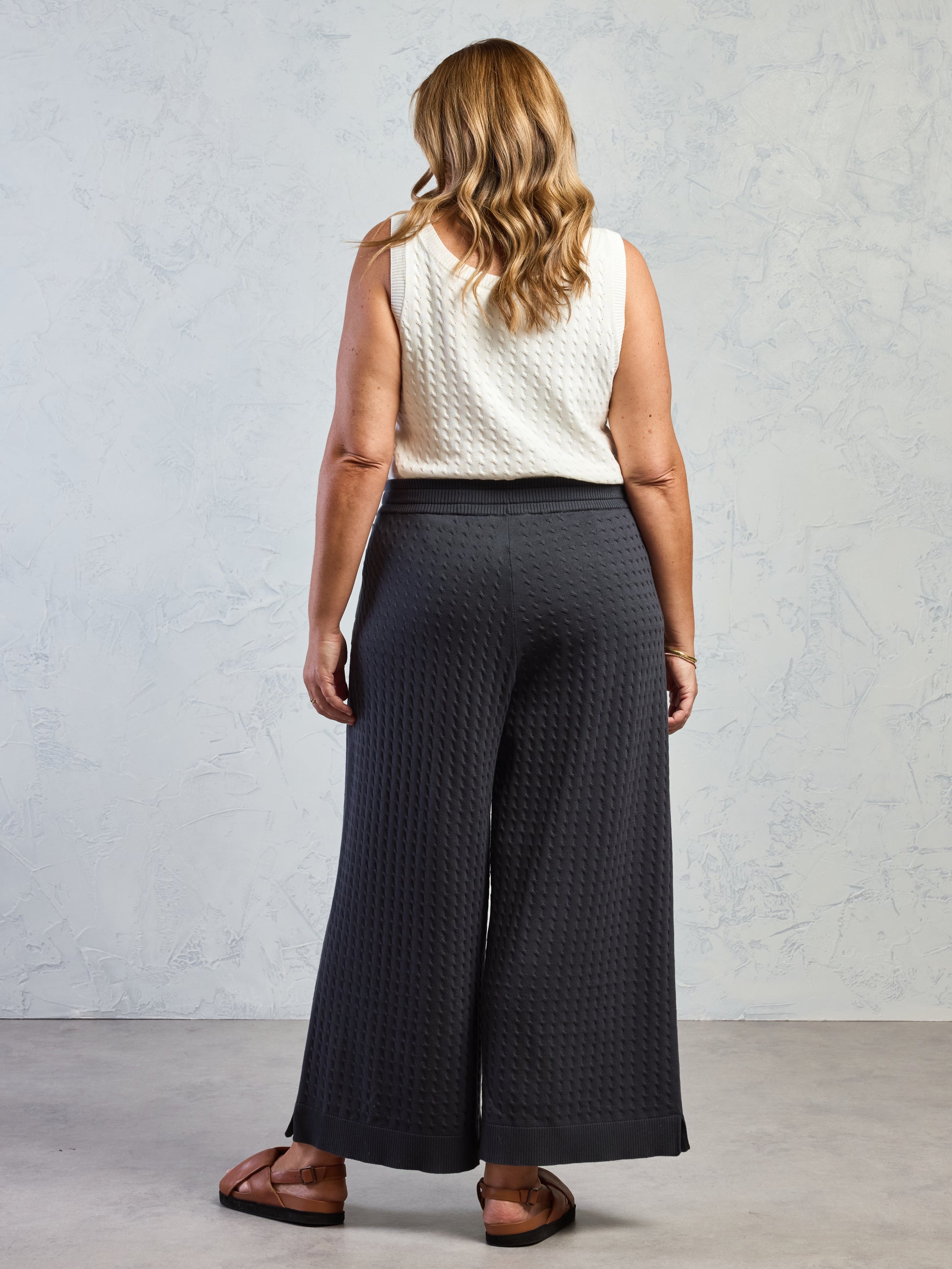 Woman wearing a white sleeveless top and dark gray wide-leg pants against a plain background