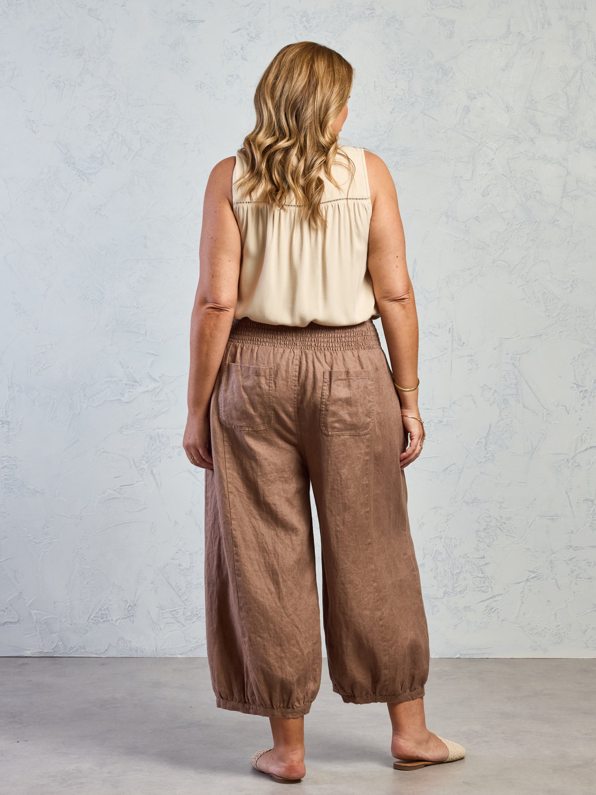 Woman wearing a beige sleeveless top and brown wide-leg pants against a light gray background