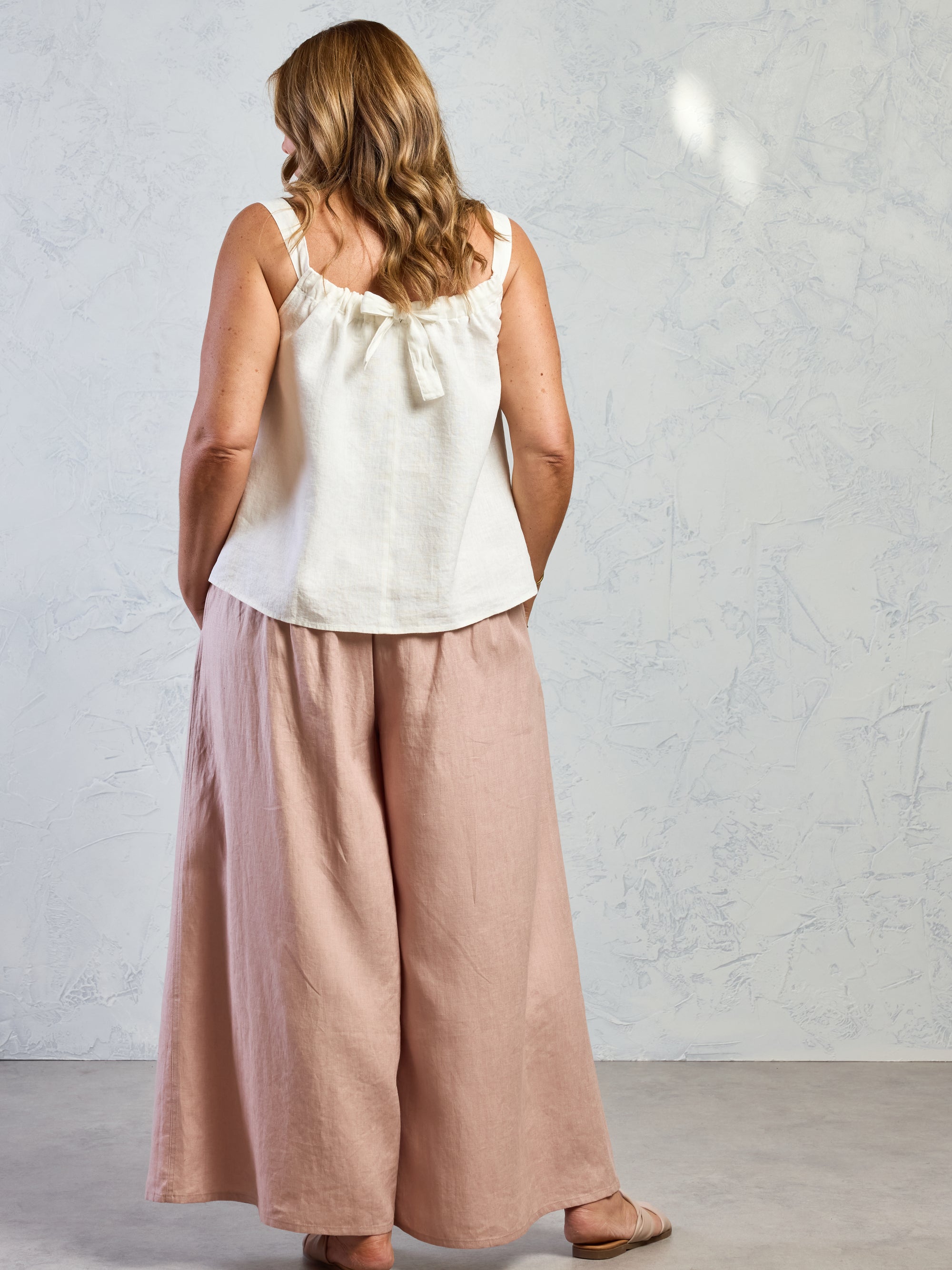 Woman wearing a white sleeveless top and pink wide-leg pants against a gray background