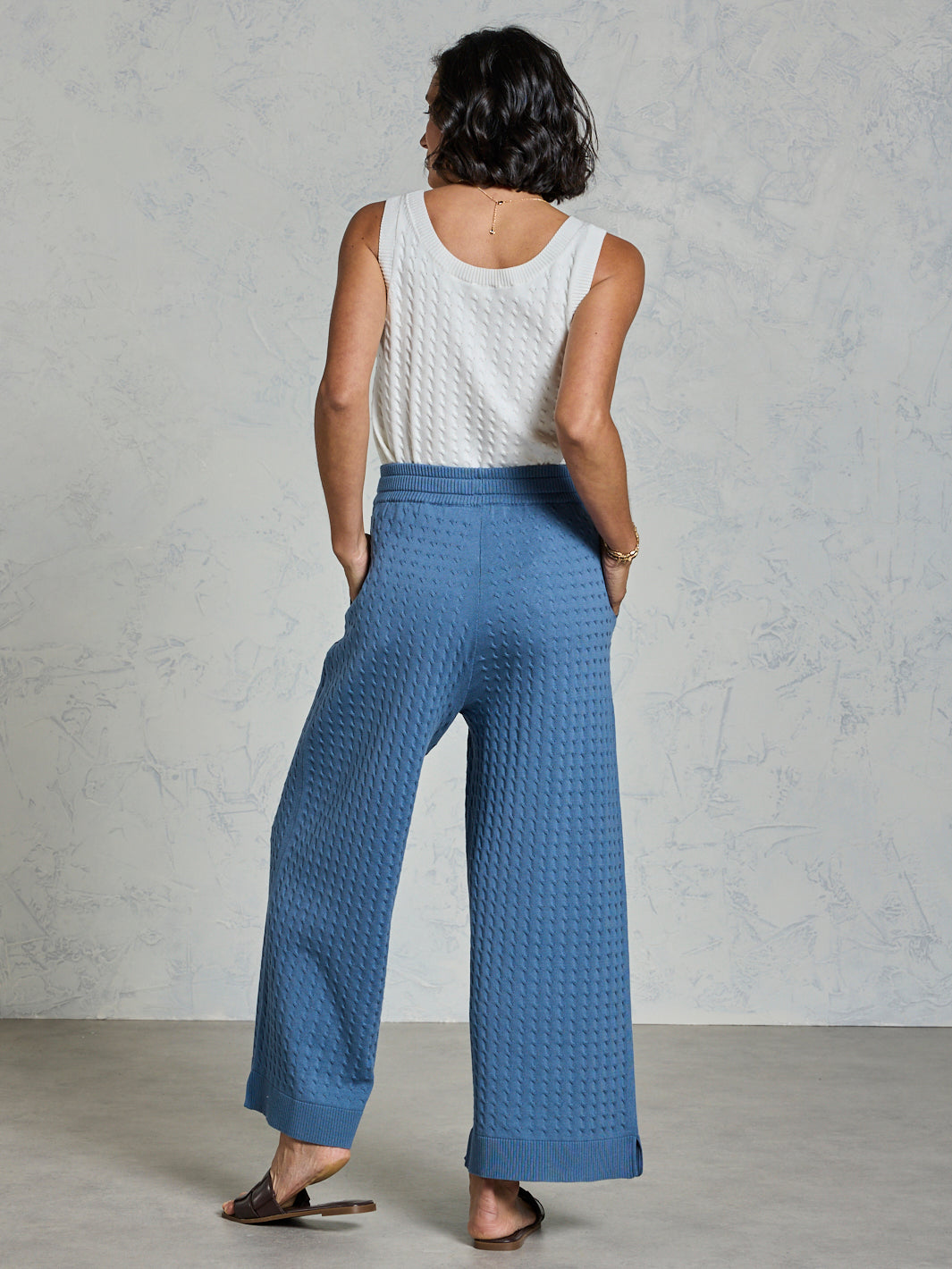 Woman wearing blue textured pants and a white sleeveless top against a gray background
