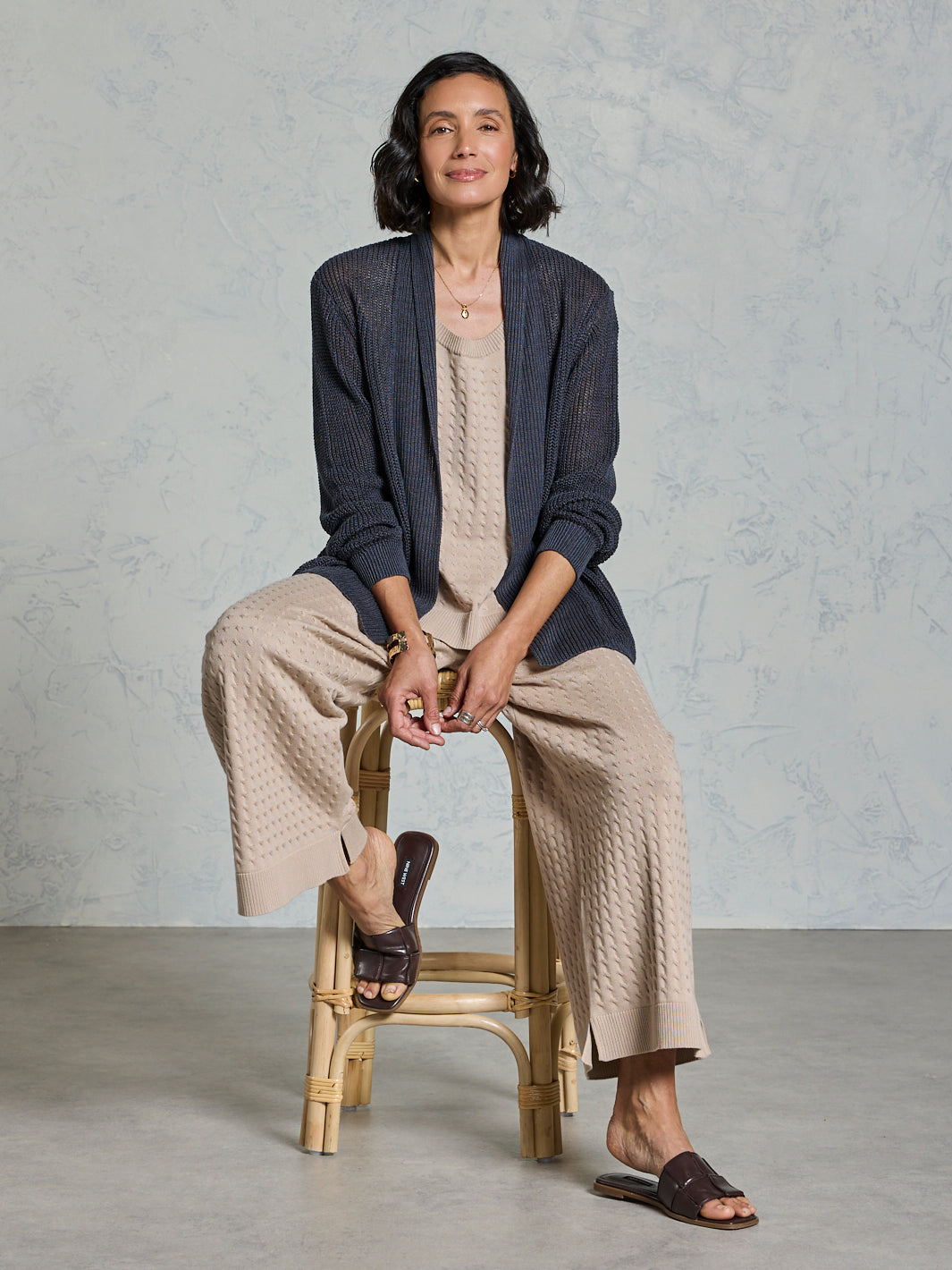Woman sitting on a stool wearing a navy cardigan and beige pants against a gray background