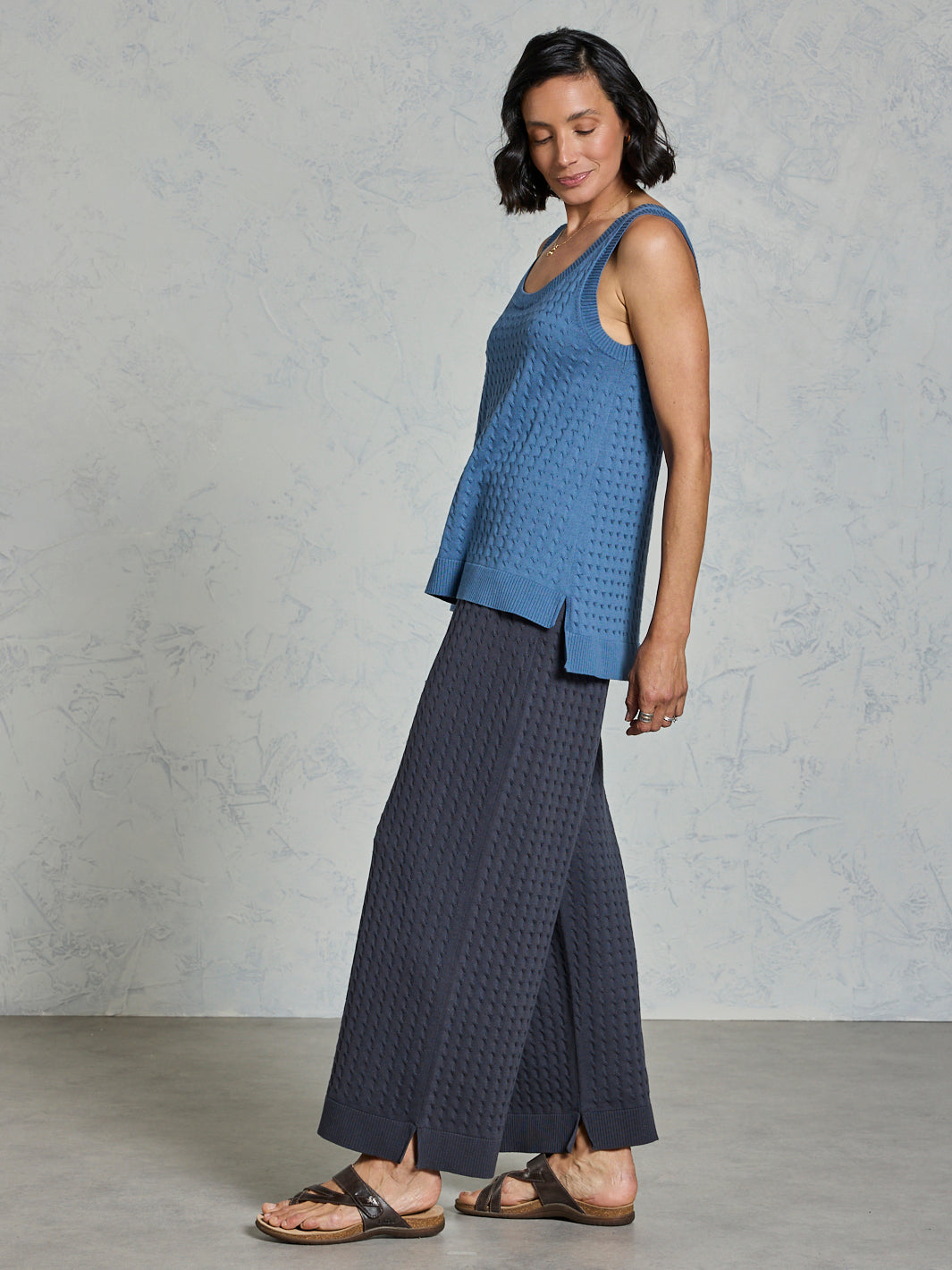 Woman wearing a blue sleeveless top and dark pants against a gray background