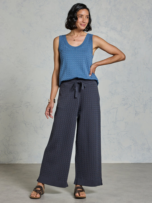 Woman wearing a blue sleeveless top and dark patterned pants against a gray background