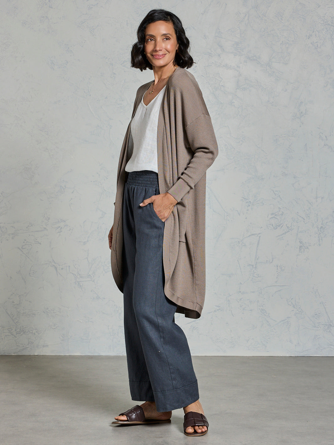 Woman wearing a long beige cardigan over a white top and dark pants against a plain background