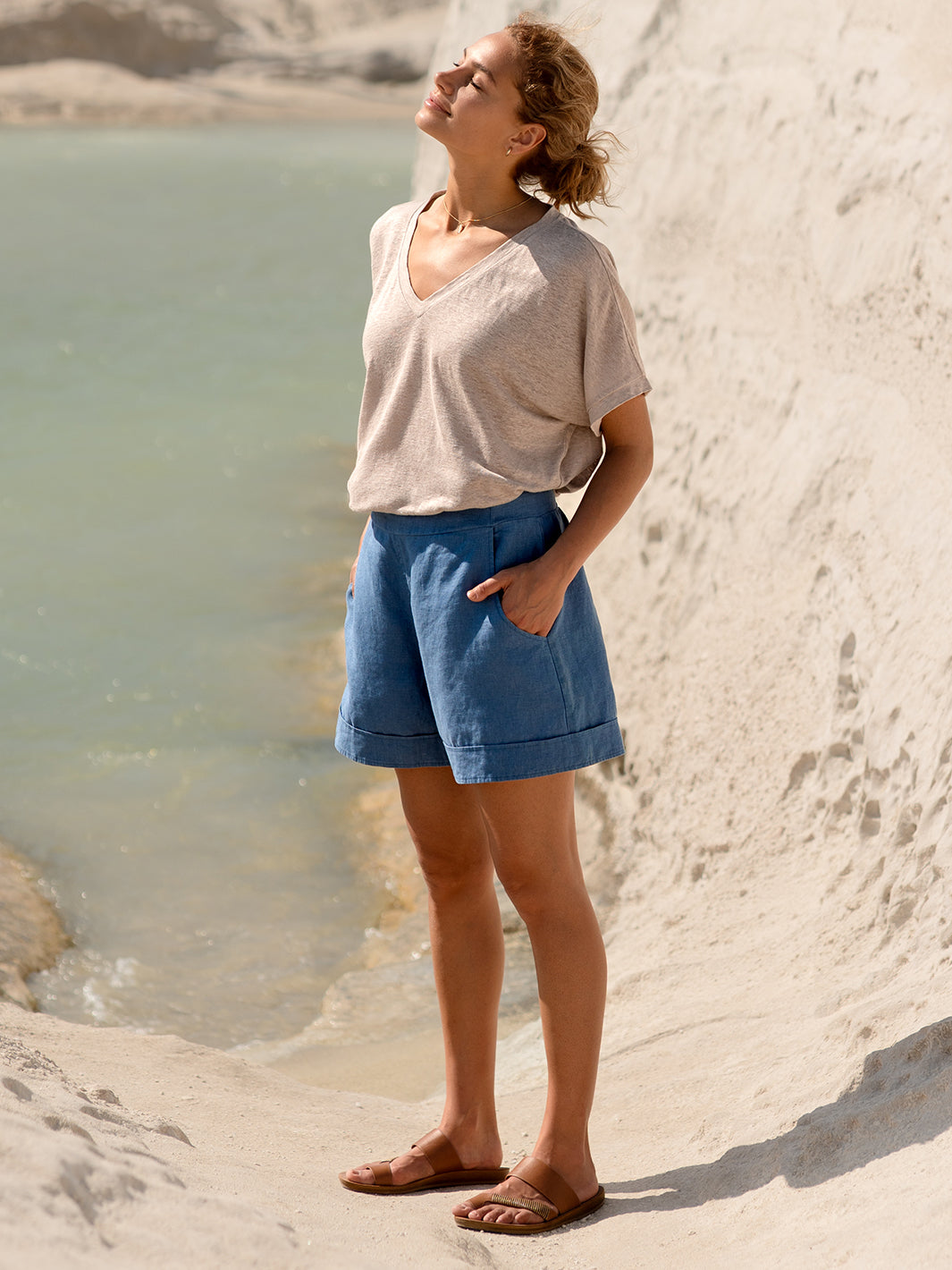 Woman standing by a body of water wearing a beige shirt and blue shorts.