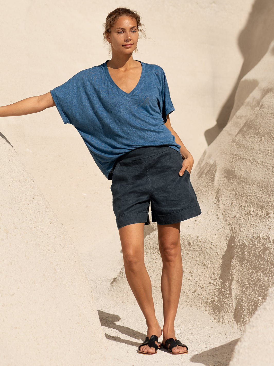 Woman wearing a blue shirt and dark shorts standing against a textured beige wall.