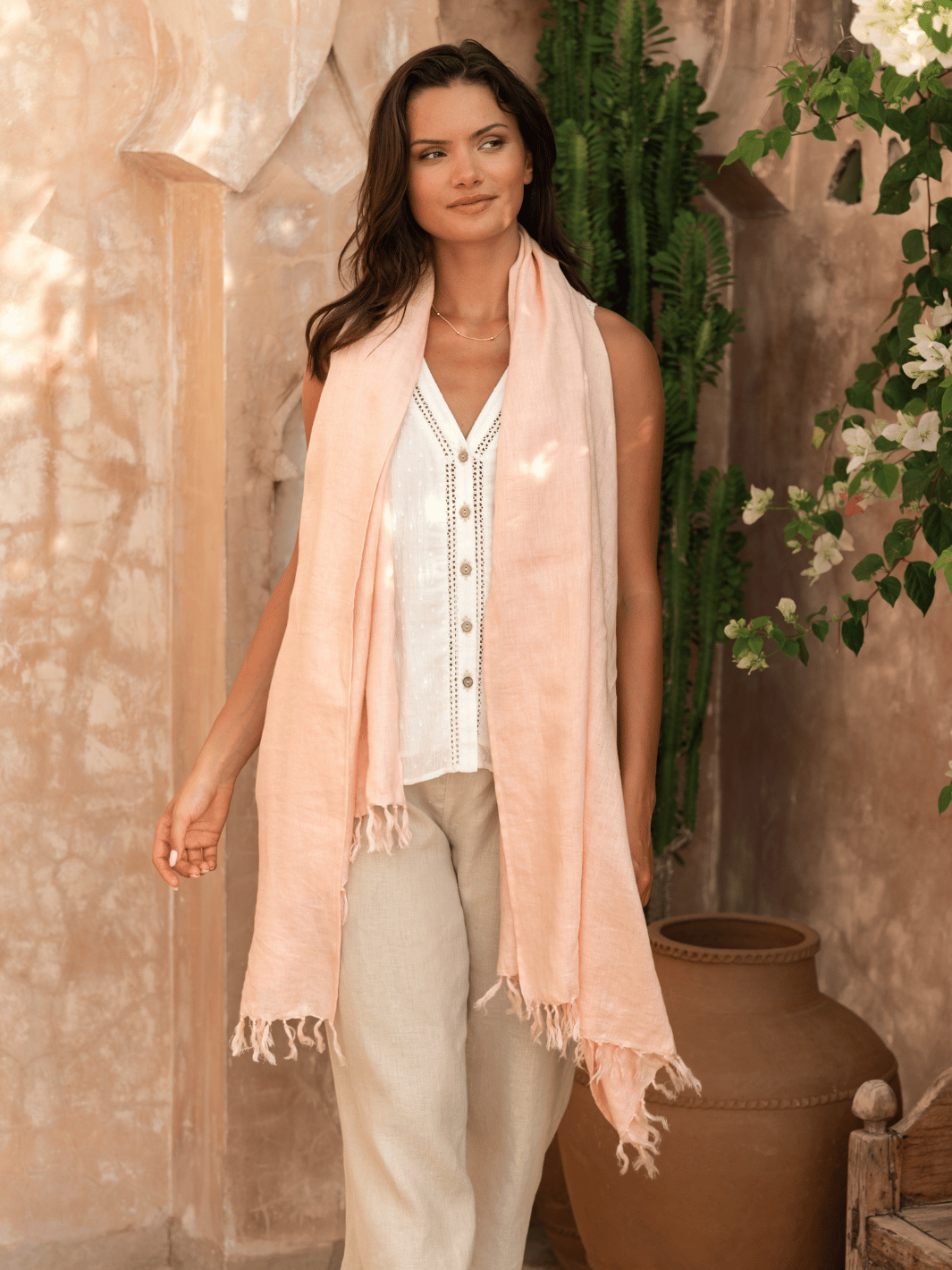 A woman wearing a coral linen scarf with tassels, paired with a white top and beige pants, standing near a pot plant.