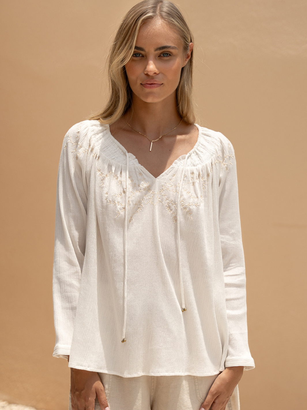 A woman wearing a white, long-sleeved gauze blouse with a V-neck, featuring smocked neckline with adjustable drawcord, open keyhole back, and signature embroidery with antique brass bead details.
