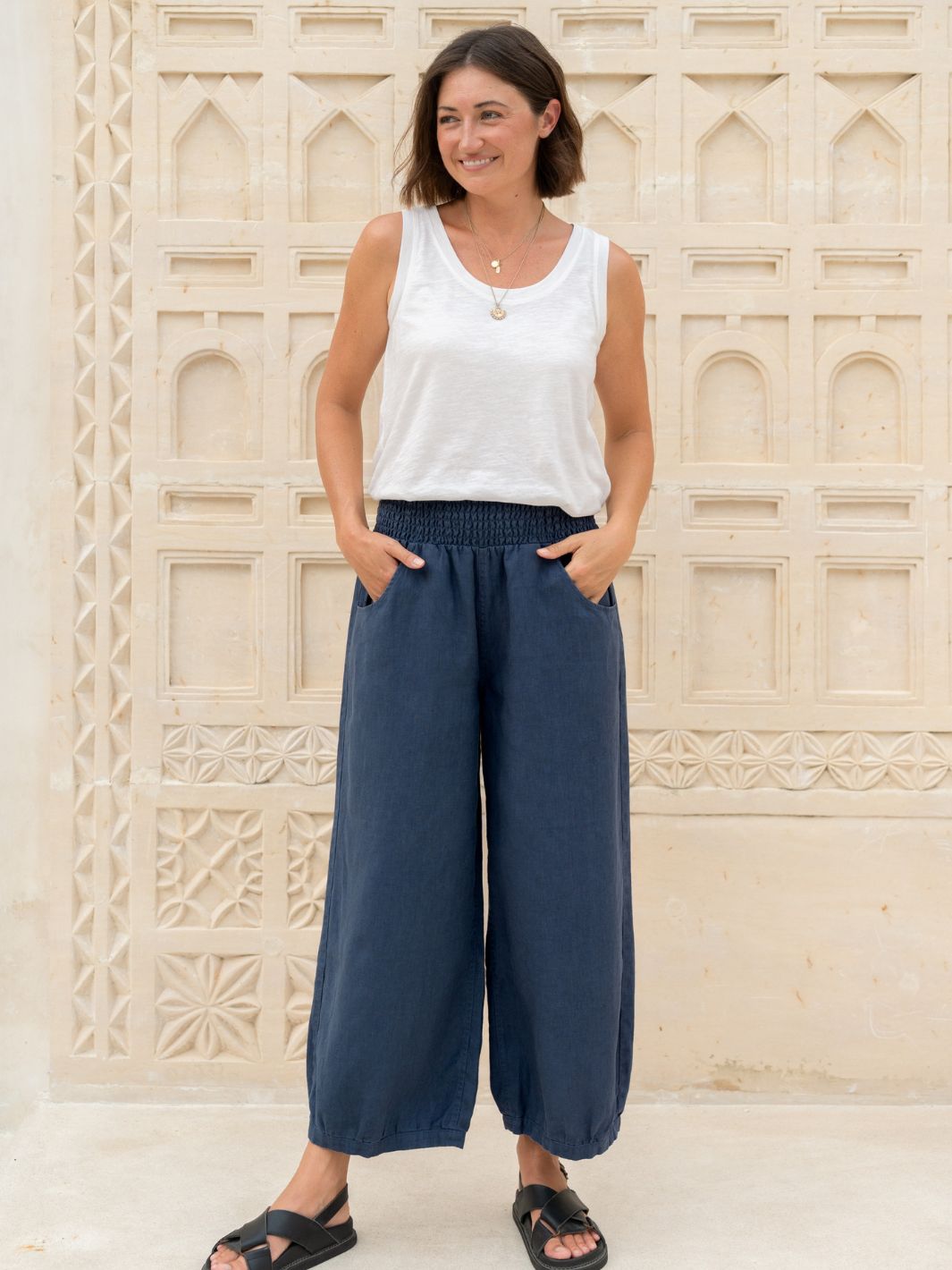 Woman wearing a white sleeveless top and blue wide-leg pants standing against a decorative stone wall.