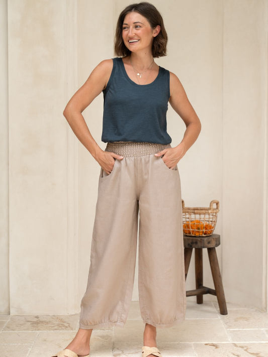 Woman wearing a black tank top and beige wide-leg pants standing in a room with a wooden stool and basket in the background.