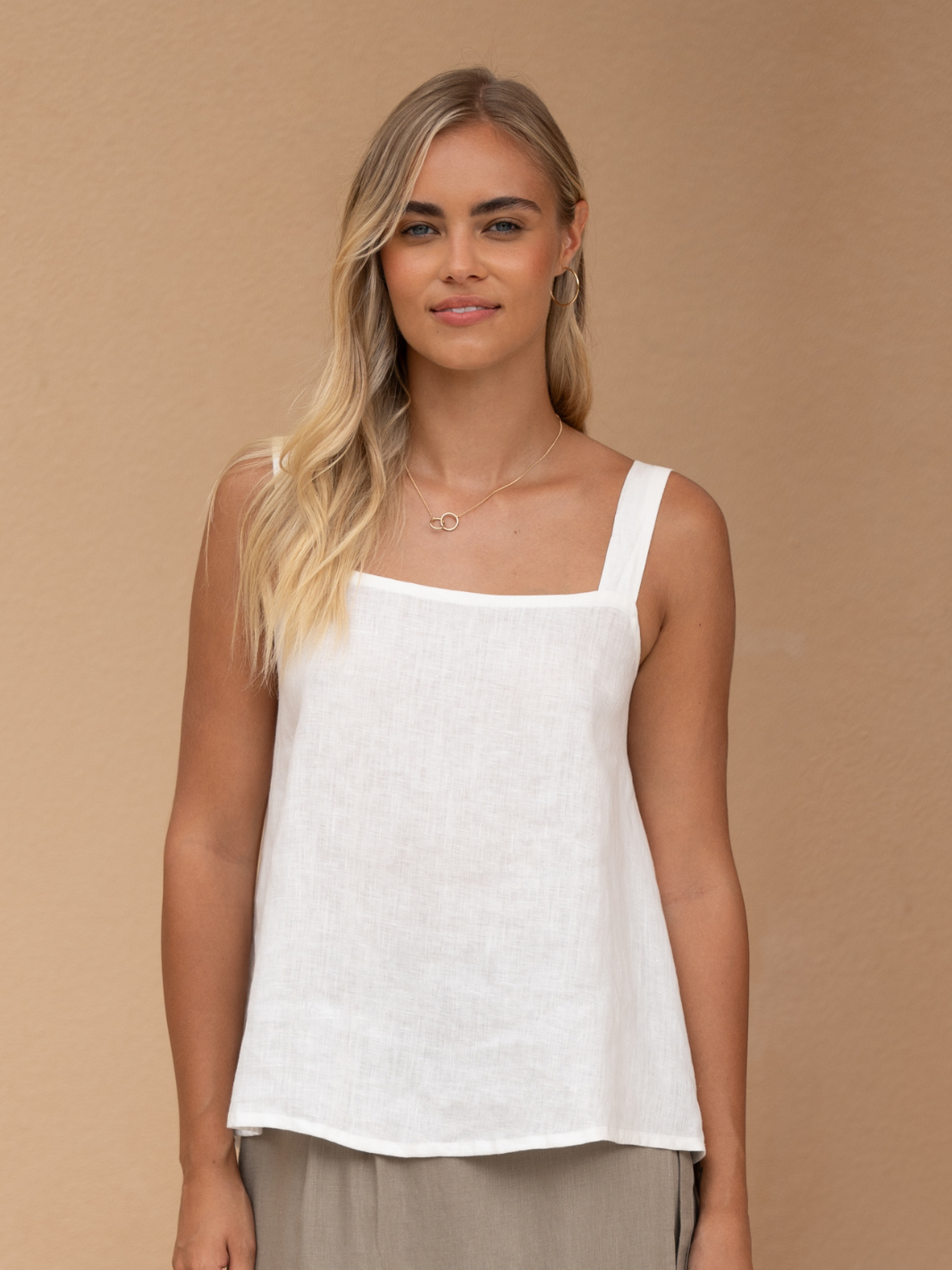 A female model wearing a white linen top with a square neckline and adjustable ties, paired with a beige skirt.