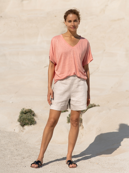 Woman wearing a pink shirt and beige shorts standing on sandy ground with a minimal background