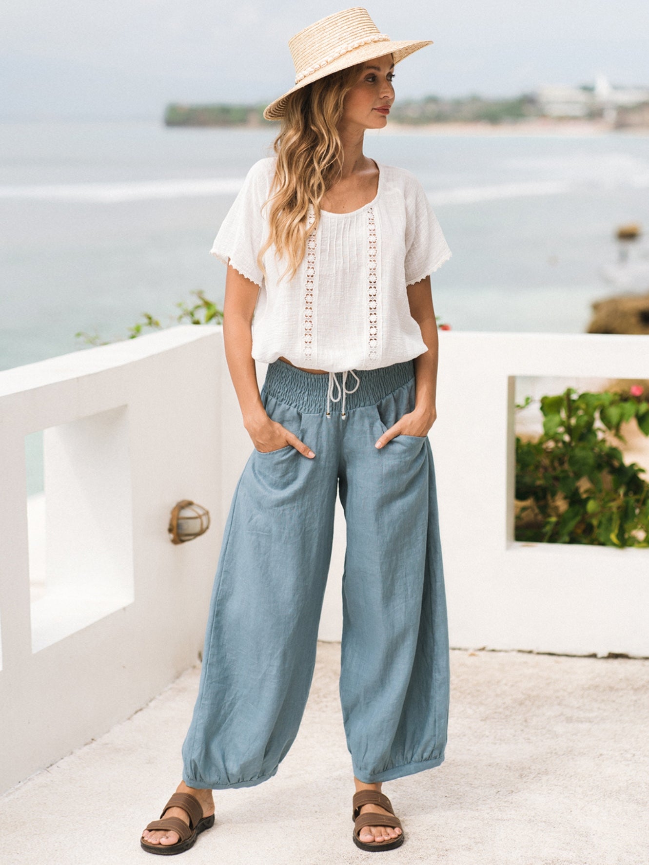 A model wearing blue linen pants with a relaxed fit and elastic waistband, paired with a white top and a straw hat.