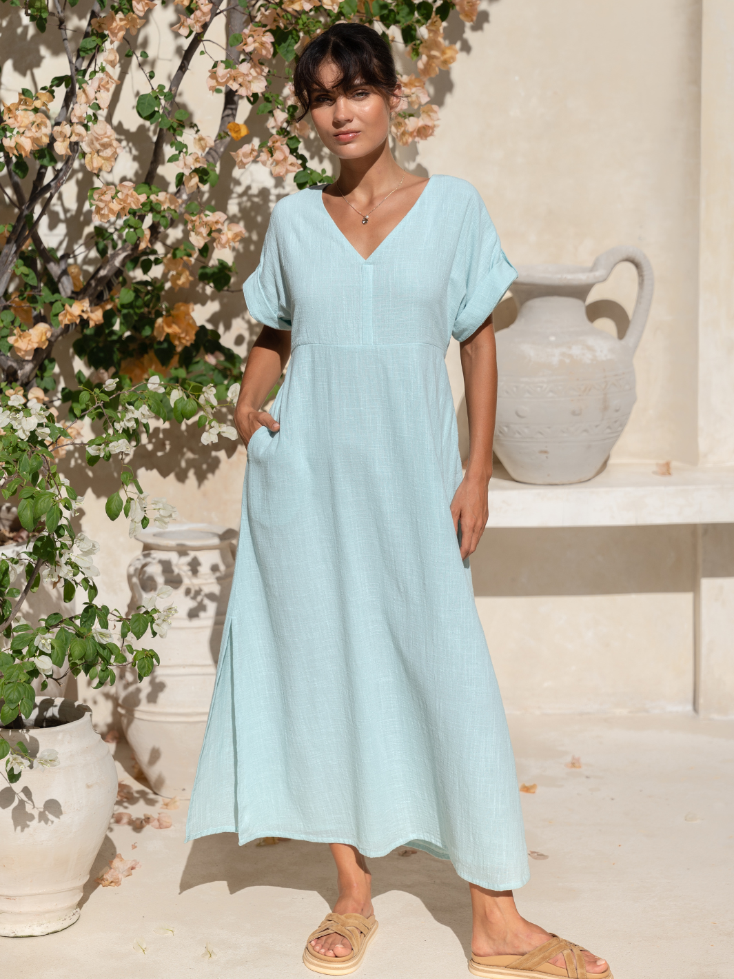 A woman wearing a seaglass blue maxi dress with a V-neckline, waist ties, and short roll-up sleeves, standing near a plant.