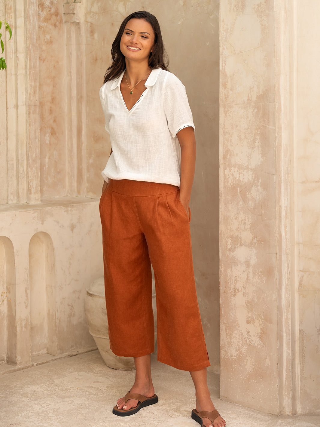 A woman standing in a relaxed pose wearing rust-colored, mid-rise, straight leg linen pants with a white top.