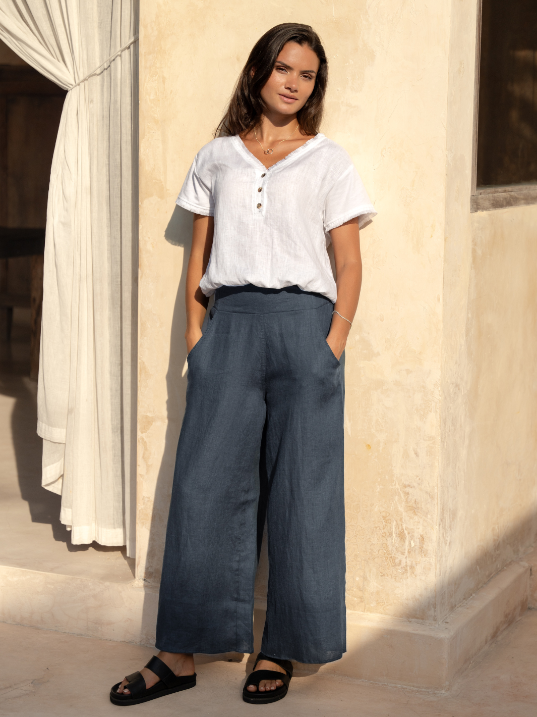 A woman standing in a charcoal gray linen wide-leg pants with a wide classic waistband, deep front pockets, and no back pockets. She is wearing a white top and black sandals, posing with her hands in the pockets.