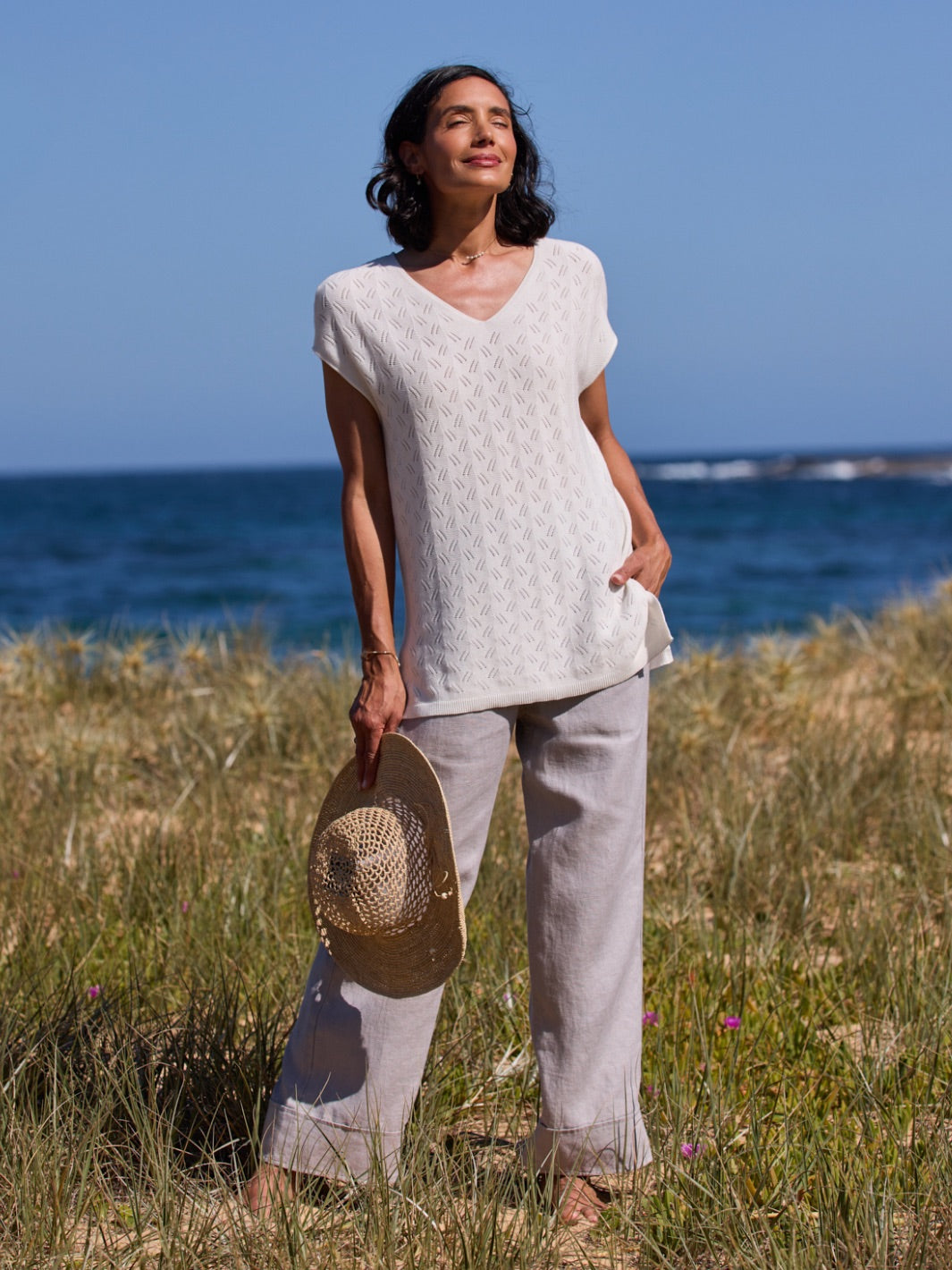 Woman standing on a grassy beach holding a hat with ocean in the background