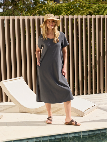 Woman in a gray dress and straw hat standing by a poolside with a lounge chair.