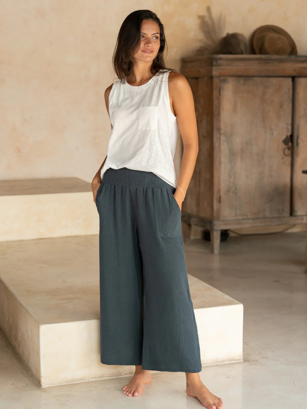Woman wearing a sleeveless top and wide-leg pants standing on stone steps with a wooden cabinet in the background.