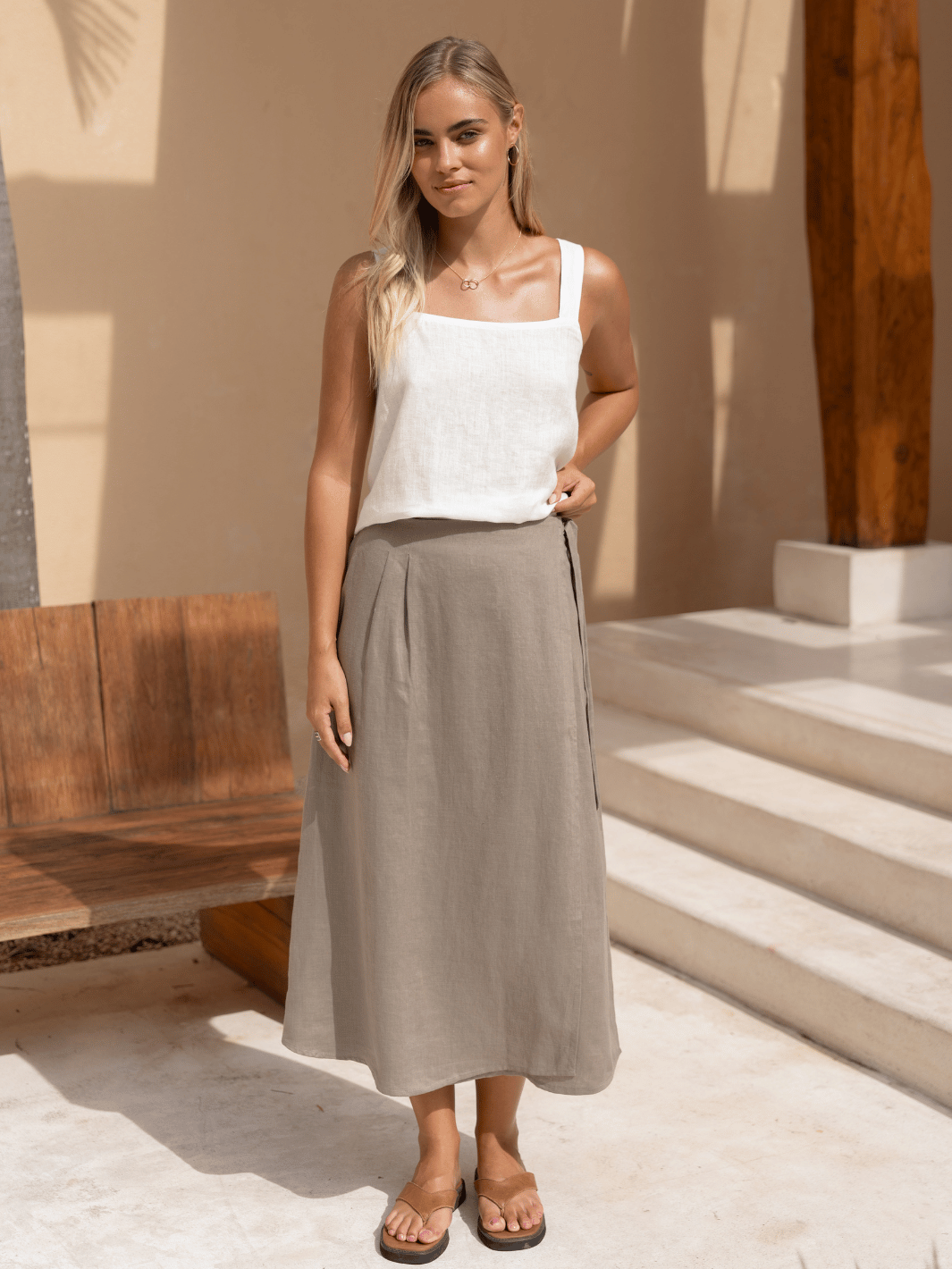 A model wearing a beige linen midi skirt with a A-line shape and a flexible waist, paired with a white tank top.