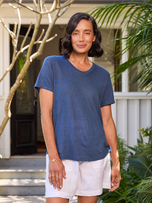 Woman wearing a blue shirt and white shorts standing outdoors with plants and a building in the background