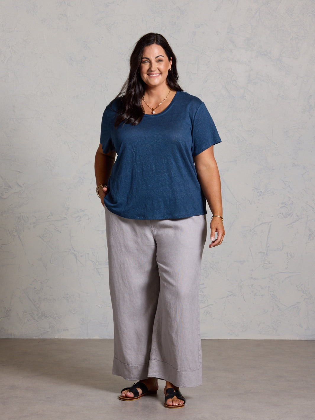 Woman wearing a blue shirt and light gray pants against a plain background