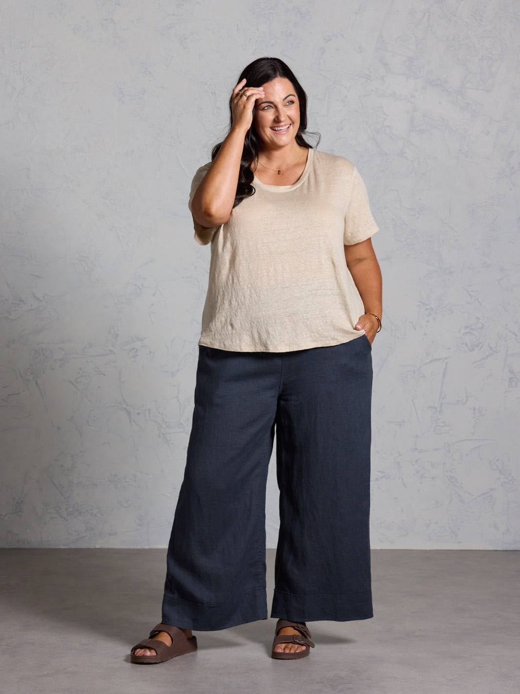 Woman wearing a beige top and black pants against a gray background