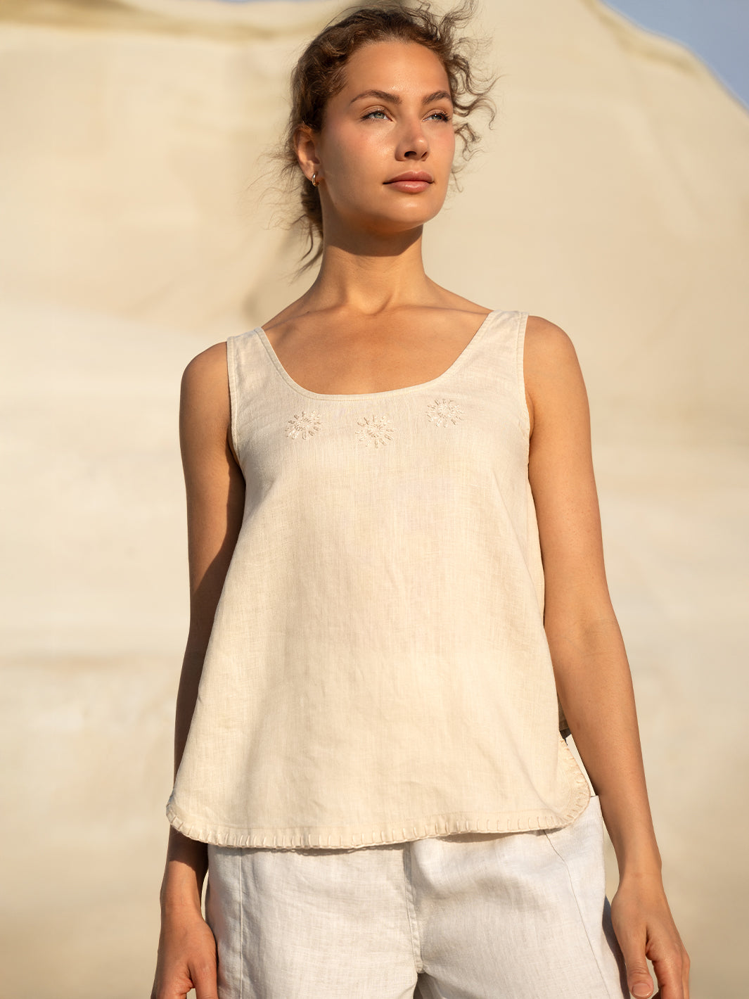 Woman wearing a beige sleeveless top and white pants against a beige background