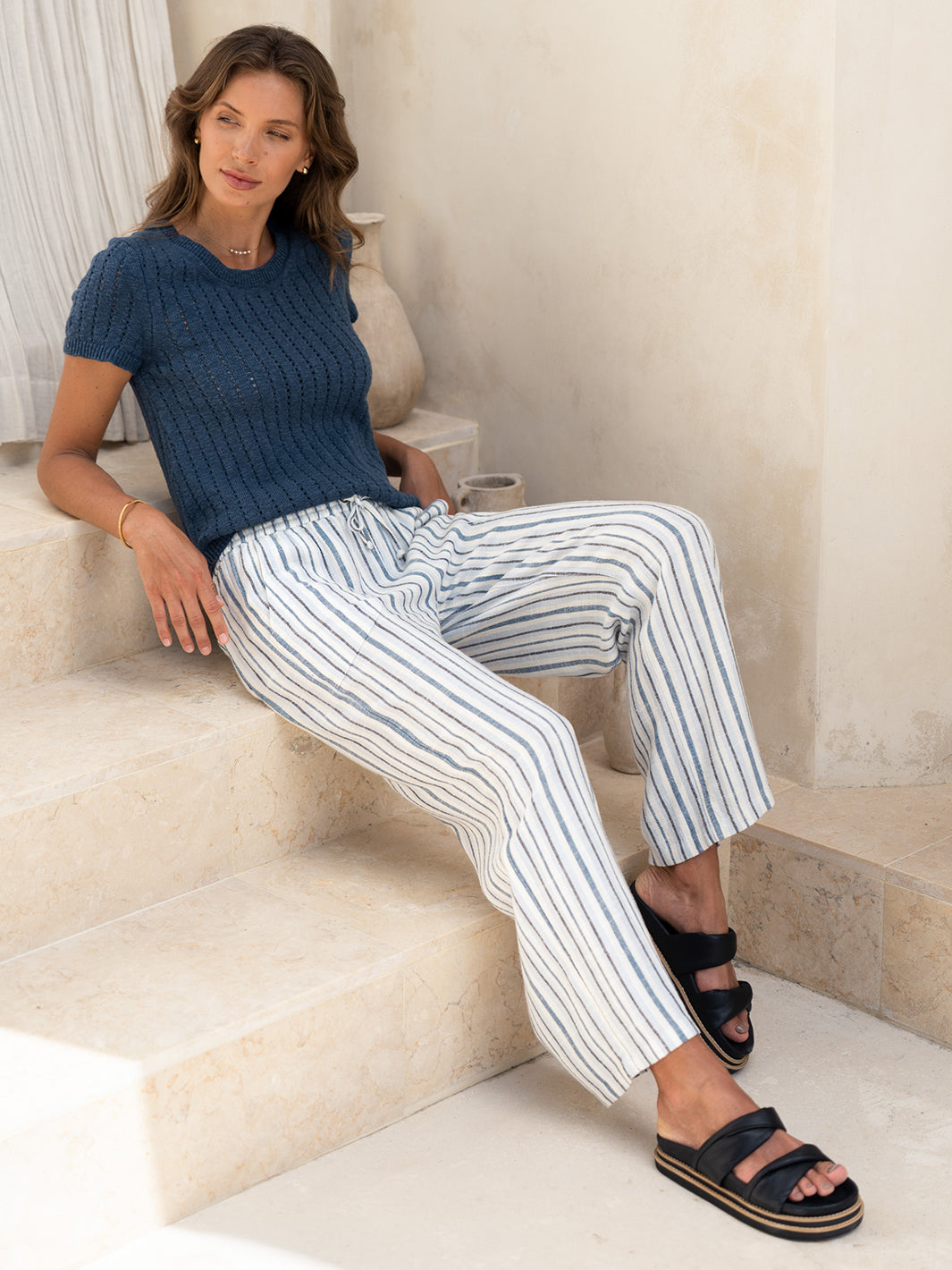Model seated in the Jolie Linen Blend Pants in coastal blue with a navy knit top, on sunlit stone steps.