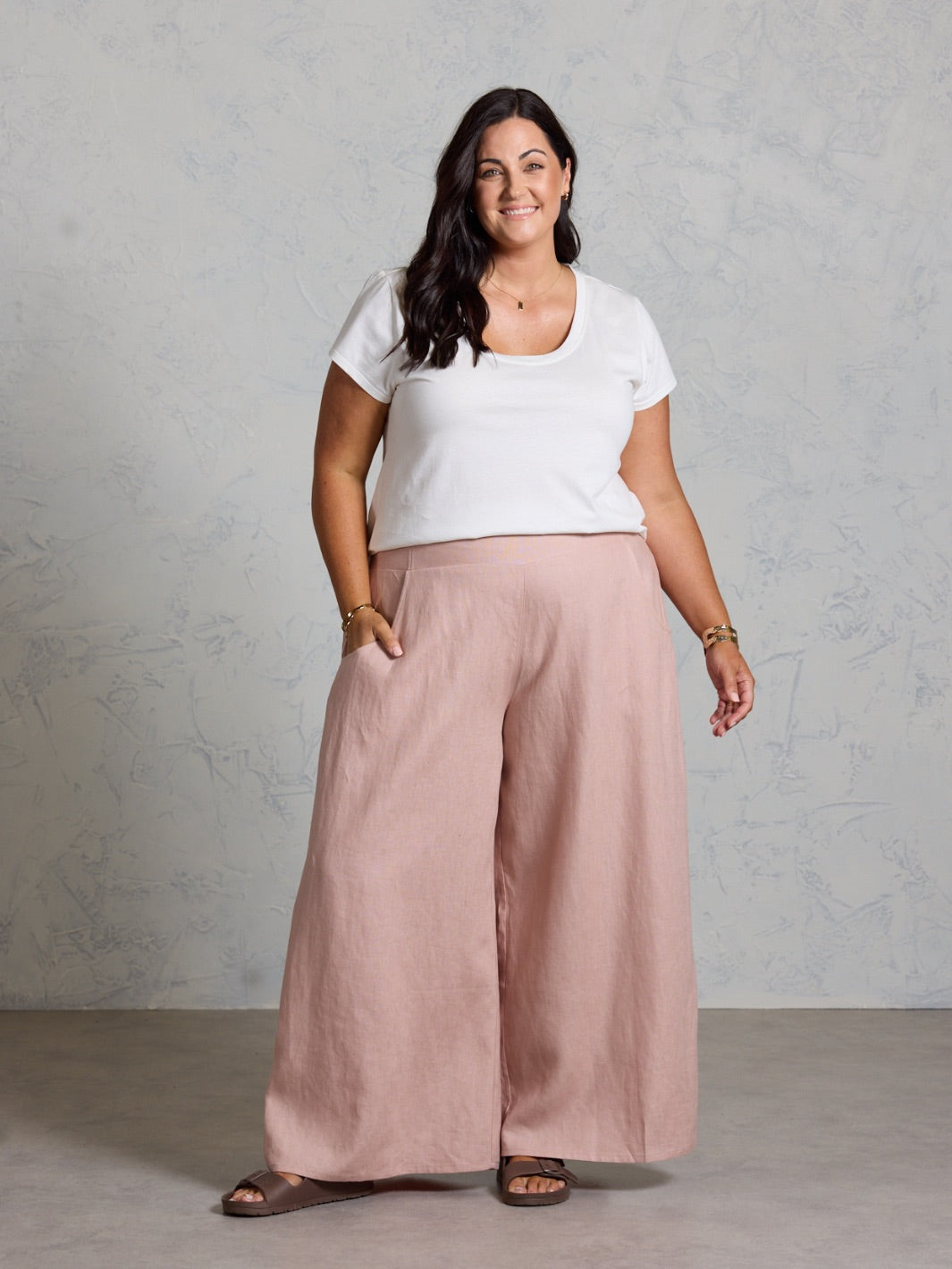 Woman wearing a white top and pink wide-leg pants against a gray background