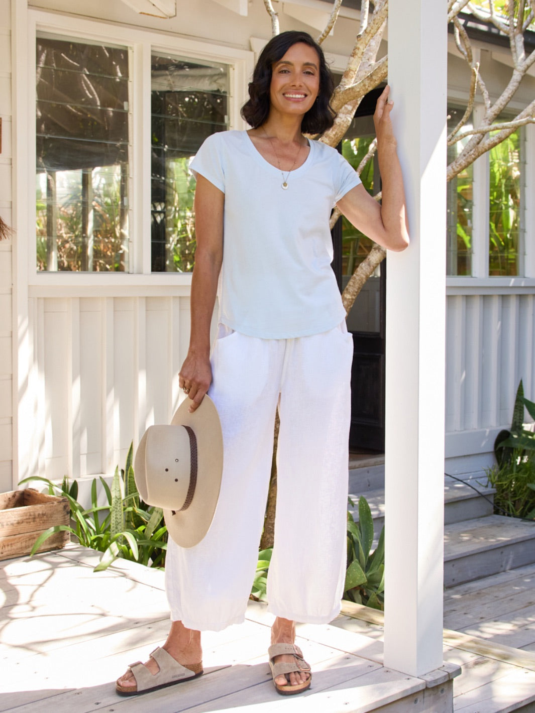 Woman in light blue top and white pants standing on a wooden deck holding a hat.