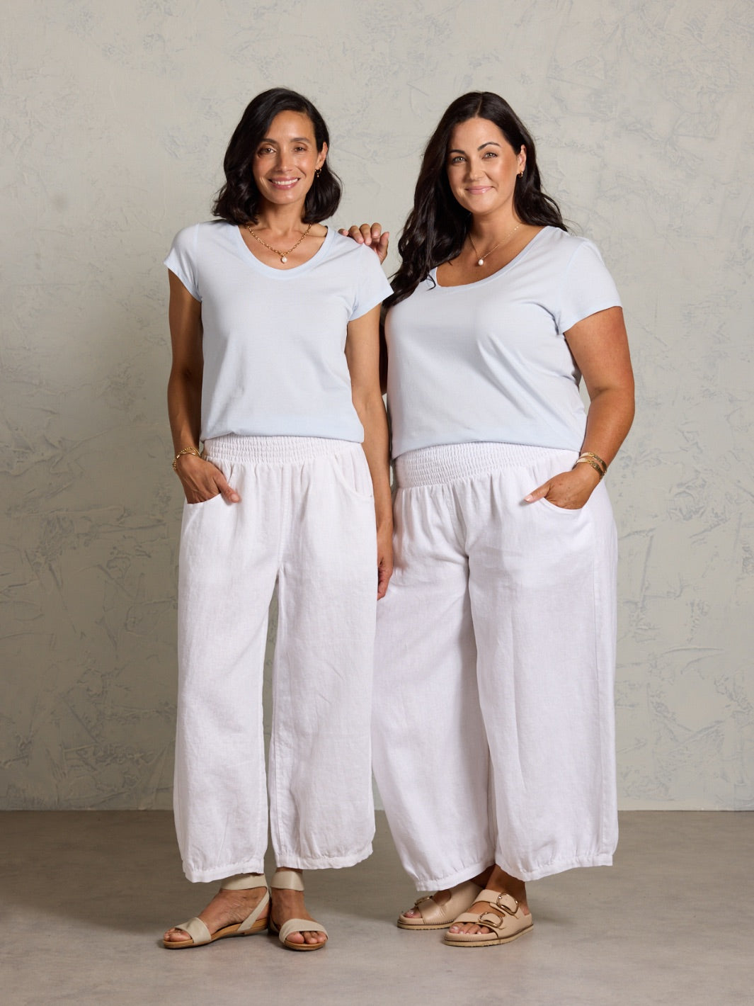 Two women wearing white tops and wide-leg pants standing against a plain background