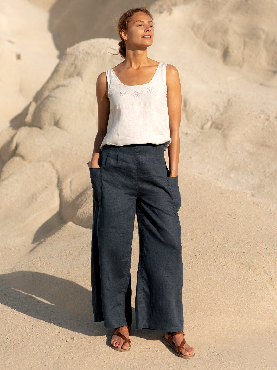 Woman wearing a white tank top and dark pants standing on sandy ground.