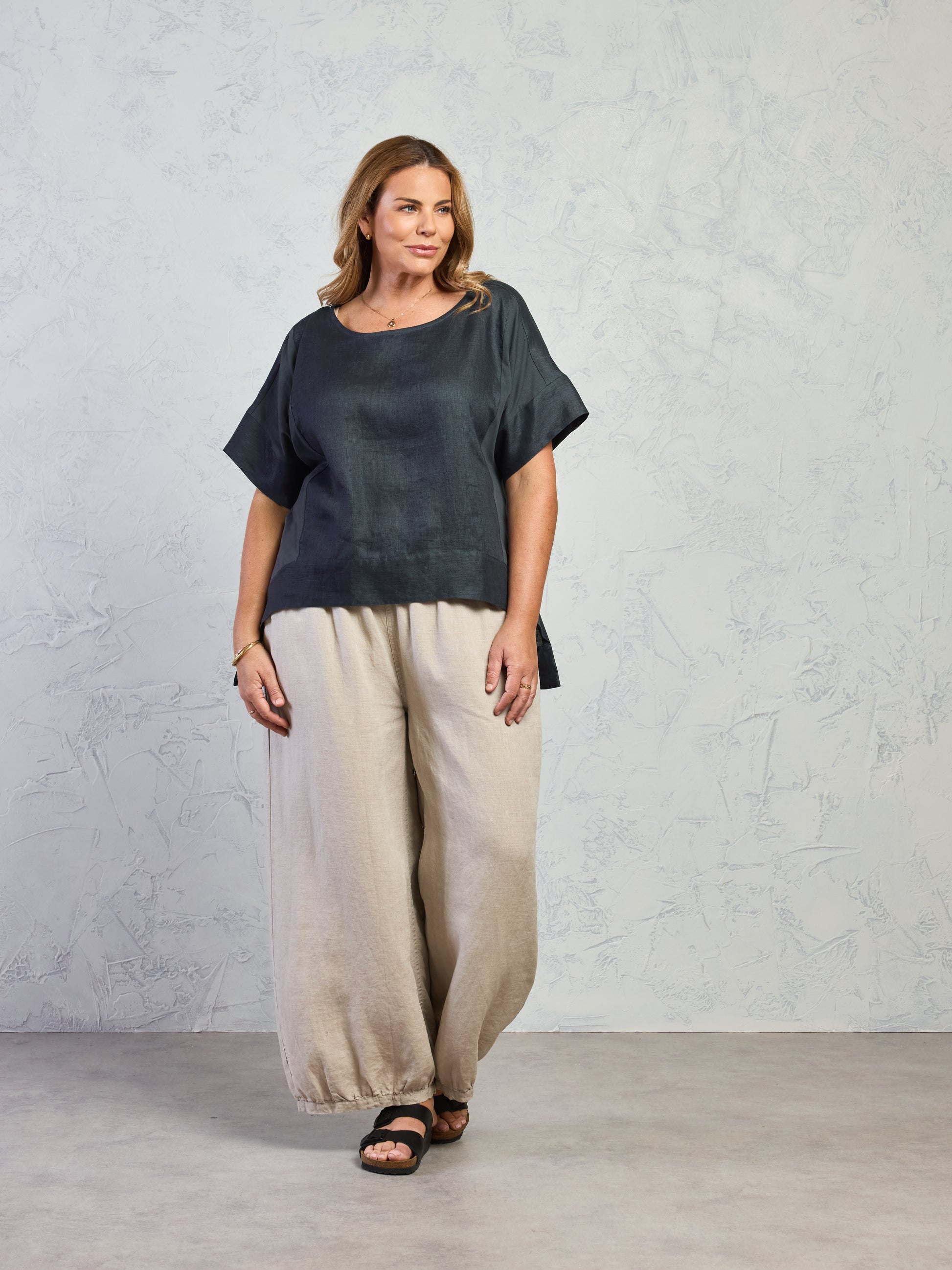 Woman wearing a dark green top and beige pants against a light gray background