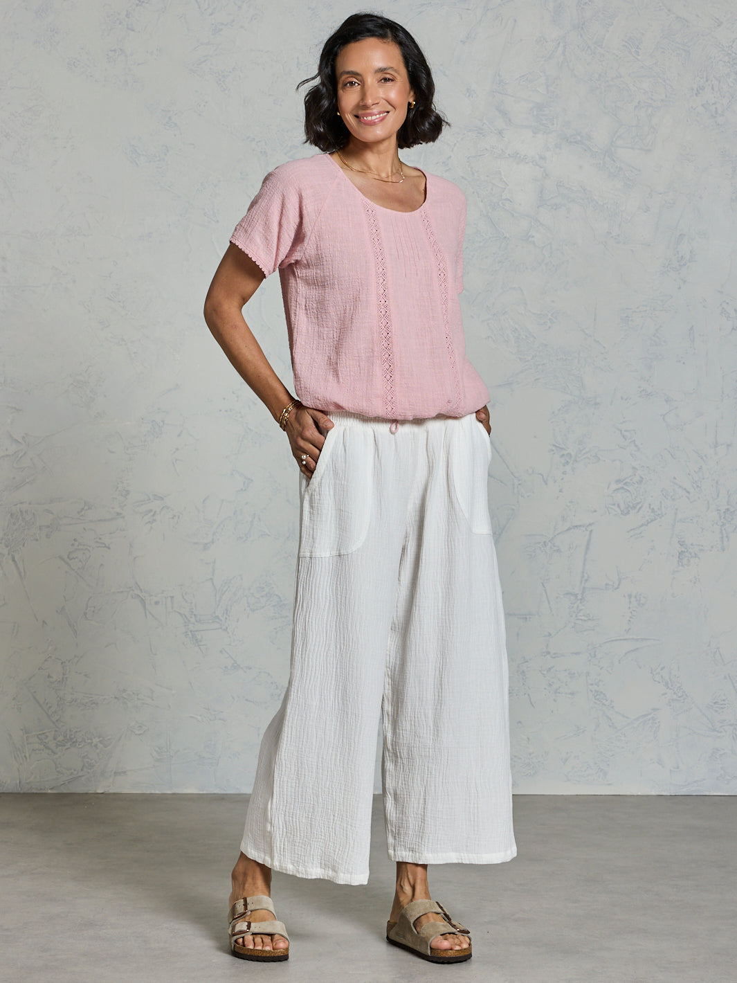 Woman wearing a pink top and white pants against a gray background